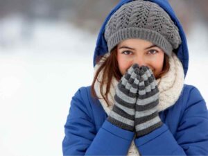 Ragazza con giubbino, sciarpa, capello e guanti che riscalda mani con il fiato della bocca